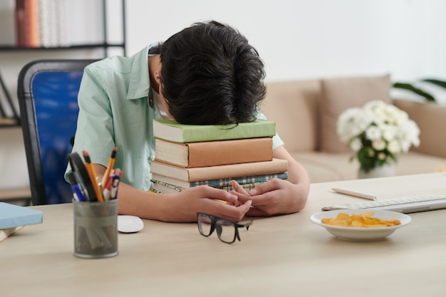 Esausto ragazzo seduto alla scrivania a casa con la testa su una pila di libri dopo aver studiato per l'esame