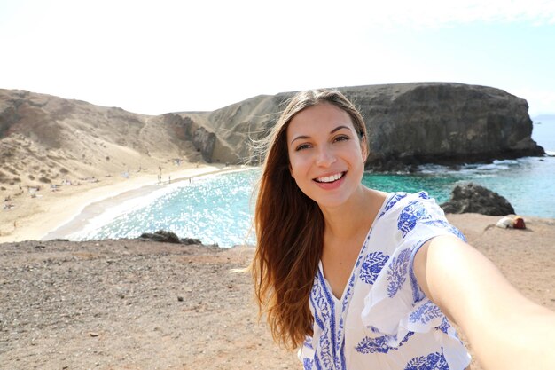 Escape Holidays Cute giovane donna sorridente con il vento tra i capelli prendendo selfie sul mare a Lanzarote Isole Canarie
