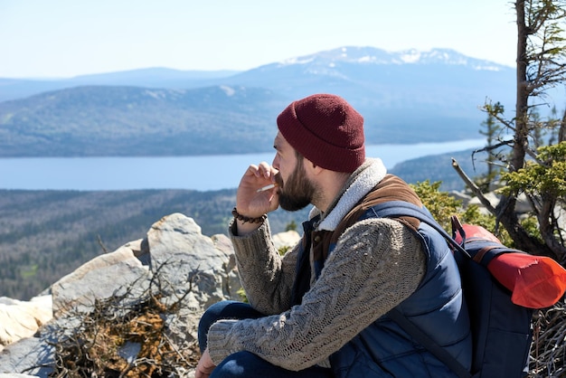 Escursioni in montagna giovane uomo barbuto con zaino appoggiato sulla cima della montagna che fuma e si gode la foto