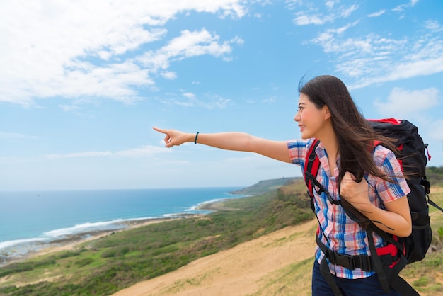 escursionista asiatico sorridente che indica il copyspace del cielo pulito condividendo lo splendido scenario con vista sull'oceano sulla cima della collina quando si viaggia in paesi insulari con la famiglia.