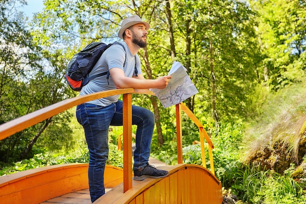 Escursionista barbuto con cappello si trova sul ponte di legno e tiene la mappa della zona dove è guidato dalla zona nella soleggiata giornata estiva.