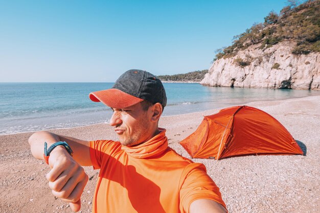 Escursionista con grimace guardando il suo orologio intelligente a portata di mano con tenda da campeggio su una spiaggia sabbiosa