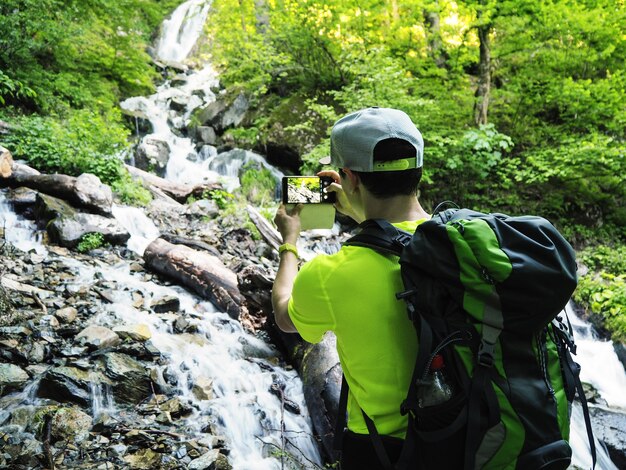 Escursionista con lo zaino che scatta foto della cascata sullo smartphone durante la soleggiata giornata estiva