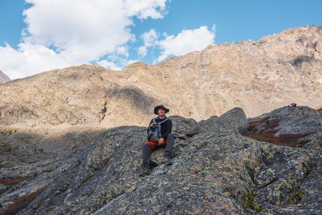 Escursionista con zaino seduto su una pietra contro le rocce alla luce del sole Solo uomo con fotocamera al sole vicino alla parete rocciosa illuminata dal sole Turista su morene in alte montagne in una giornata di sole