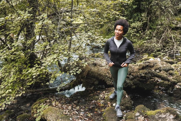 Escursionista donna afroamericana matura che attraversa un ruscello mentre esplora le montagne e si gode l'allenamento all'aperto.