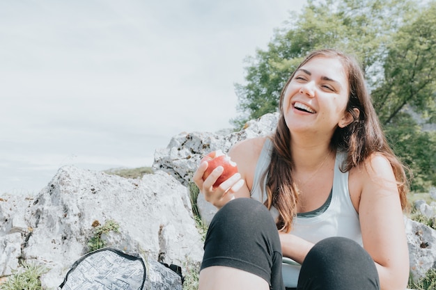 Escursionista donna che riposa e mangia una mela seduta mentre ride e sorride, copia spazio, escursionismo e concetto di libertà