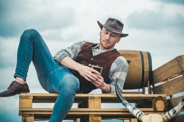 Esempio di vera mascolinità. Cappello da portare del cowboy. Vita occidentale. Ragazzo con la barba lunga in cappello da cowboy e camicia a quadri che beve alcolici. Uomo macho barbuto bello. Standard di bellezza maschile.