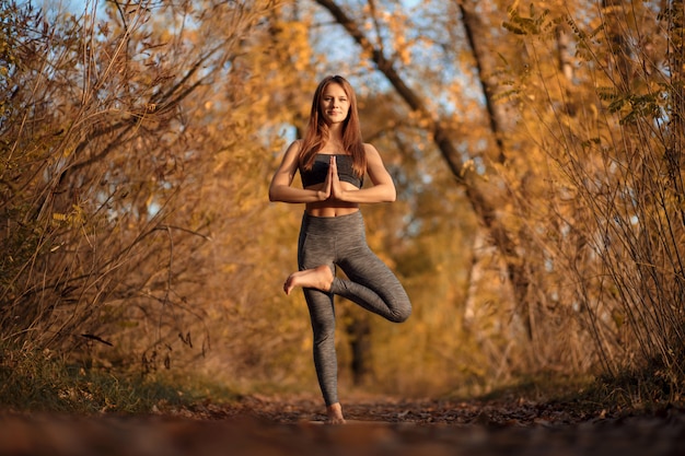 Esercizio di pratica di yoga della giovane donna al parco di autunno con le foglie gialle. Stile di vita sportivo e ricreativo