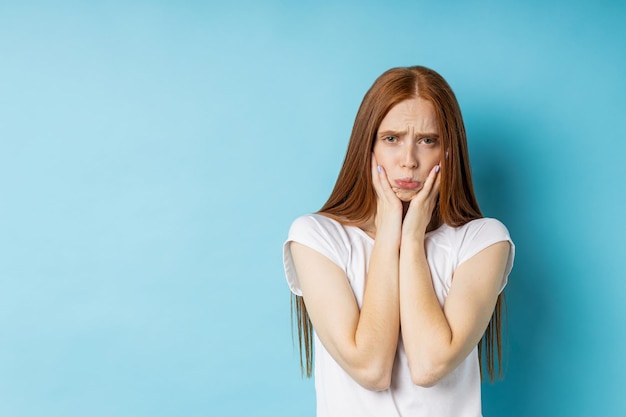 Esitante ragazza incerta con lunghi capelli rossi che allargano i palmi con espressione confusa, indossa una maglietta bianca, viso accigliato, labbra increspate, non può scegliere tra due elementi isolati su sfondo blu