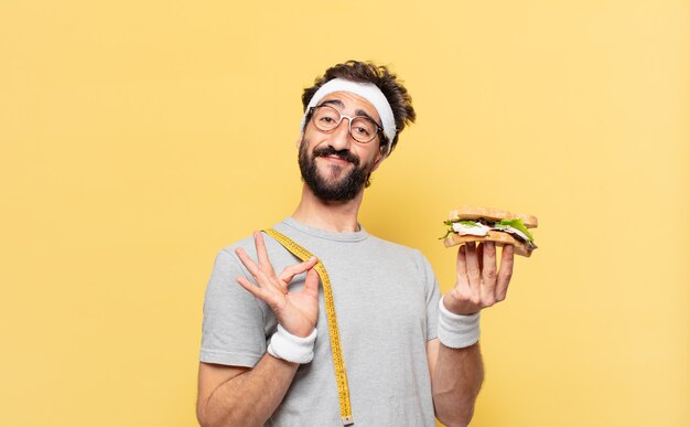 Espressione felice del giovane atleta barbuto pazzo e che tiene un panino