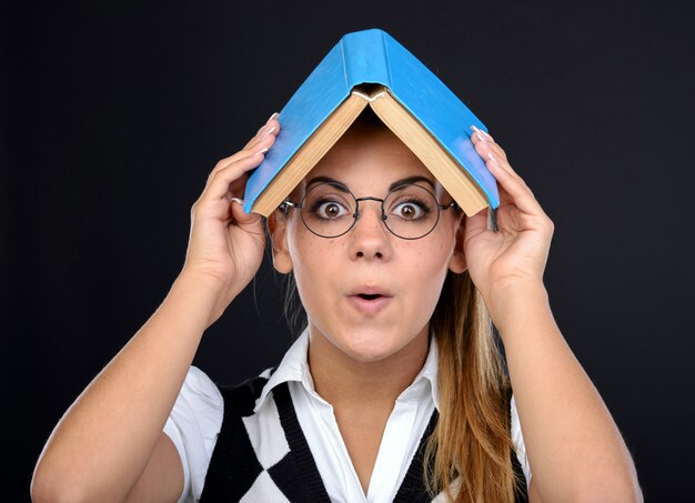 Espressione pazza della giovane donna del nerd in vetri con il libro sulla testa