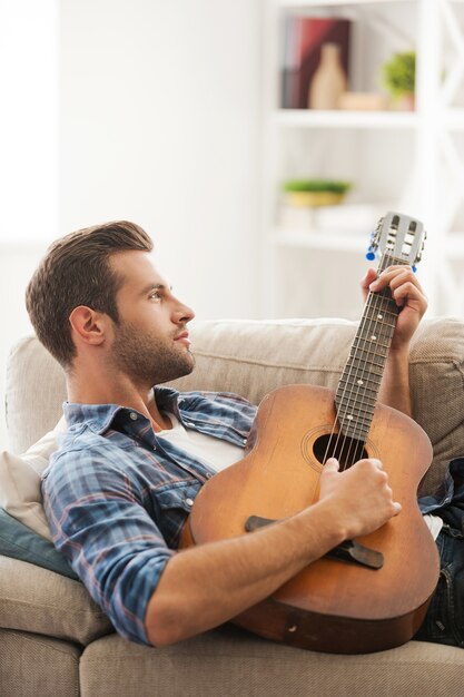 Esprimersi con la musica. Giovane premuroso che suona la chitarra mentre è sdraiato sul divano