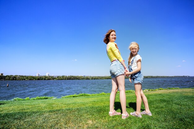 Estate e vacanza, sorelle sorridenti felici delle ragazze sul prato vicino al fiume