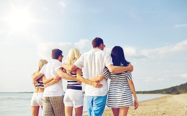 estate, vacanze, mare, turismo e concetto di persone - gruppo di amici sorridenti che si abbracciano e camminano sulla spiaggia dal retro