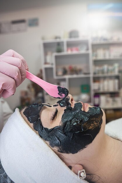Estetista che applica maschera nera al viso di una giovane donna graziosa nel salone spa