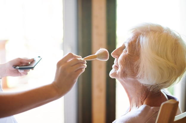 Estetista contemporanea con pennello per cipria che lavora sul trucco della signora senior prima del servizio fotografico