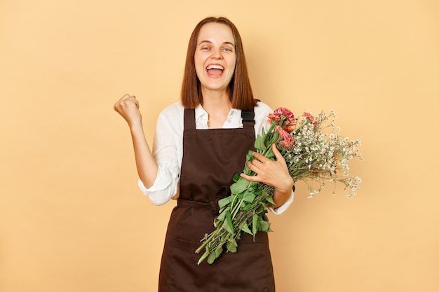 Estremamente felice sorridente allegra donna fiorista indossando un grembiule marrone tenendo un bouquet di fiori in piedi isolato su uno sfondo beige pugni chiusi celebrando il suo successo commerciale