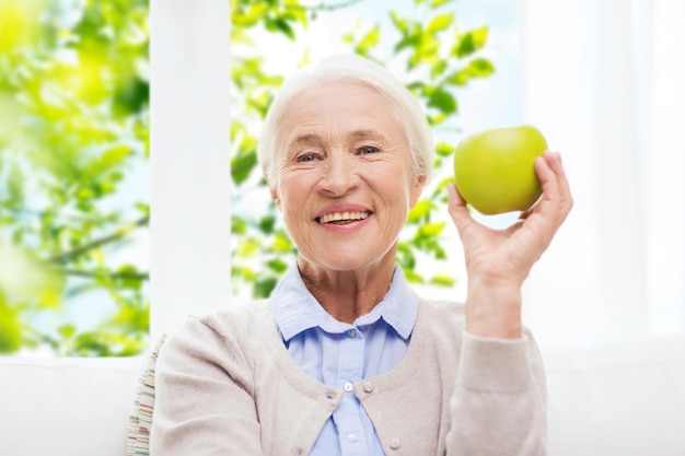 età, alimentazione sana e concetto di persone - donna senior sorridente felice con mela verde a casa sopra la finestra e sfondo verde naturale