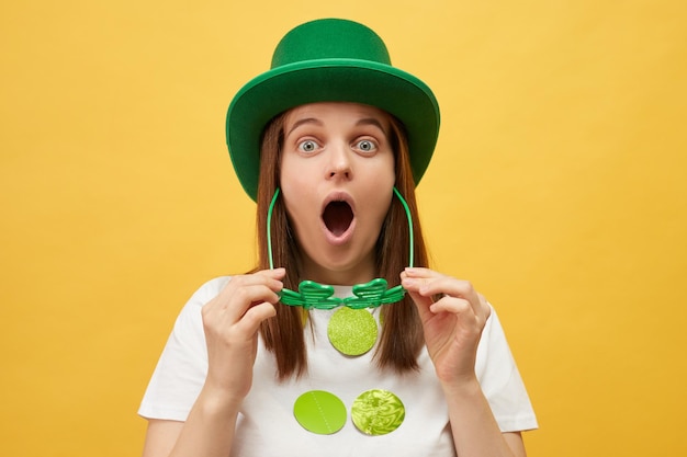 Evento a tema Shamrock Celebrazione della religione irlandese Donna scioccata che indossa un cappello verde festivo con gli occhiali di Shamrock vede qualcosa di sorprendente in piedi isolata su uno sfondo giallo