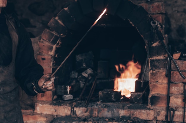 Fabbro che tiene una spada fusa incandescente vicino al forno di mattoni nella fucina Maniscalco in abiti da lavoro sporchi che lavorano il ferro