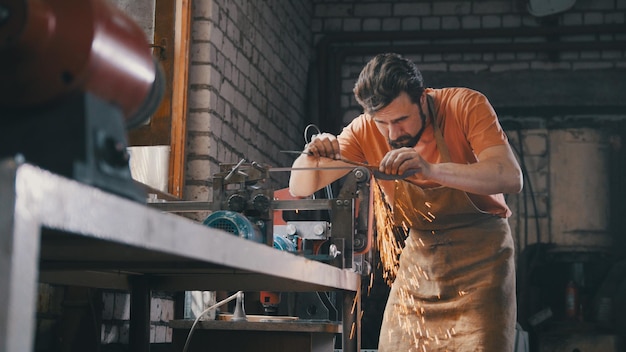 Fabbro con barba e abiti da lavoro fa una molatura di un prodotto in metallo