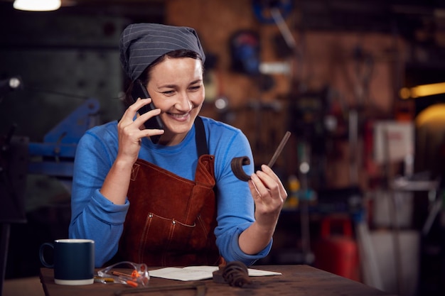 Fabbro femminile che lavora sul design in fucina mentre si utilizza il telefono cellulare