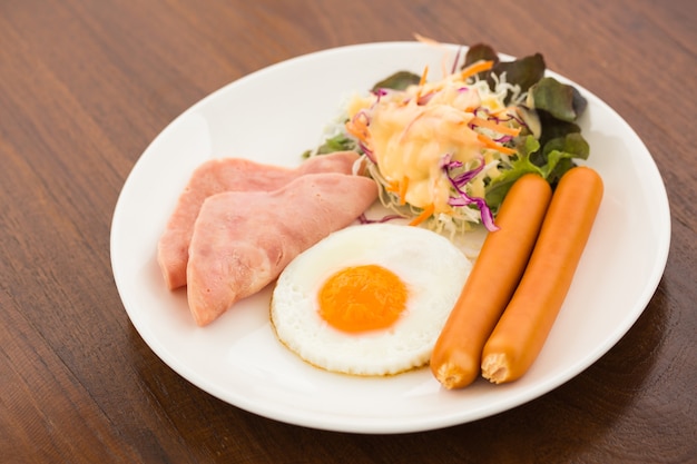 Faccia colazione con l&#39;uovo, il prosciutto, la salsiccia in piatto bianco sulla tavola di legno