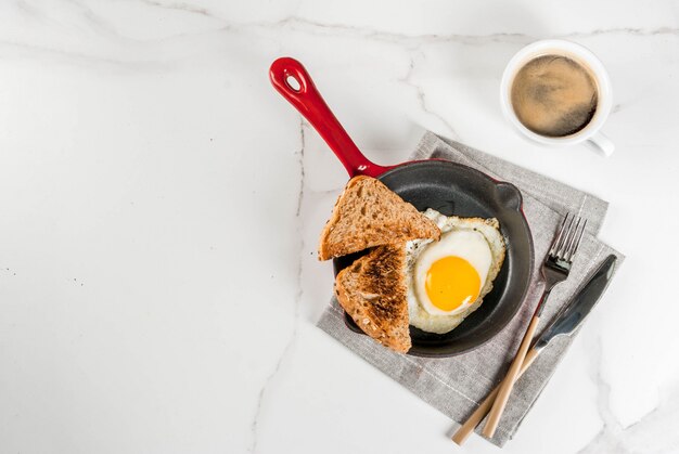 Faccia colazione con pane tostato, l'uovo fritto sulla pentola del ferro e il caffè sulla scena di marmo bianca