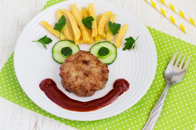 Faccia di cibo divertente con una braciola, patatine fritte e cetriolo