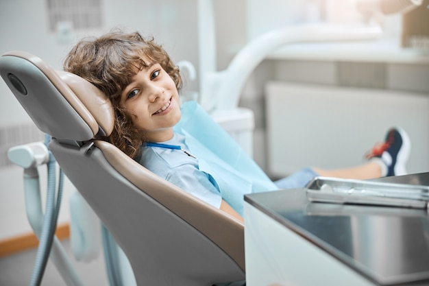 Faccina sorridente bambino dai capelli castani seduto su una poltrona del dentista guardando la telecamera