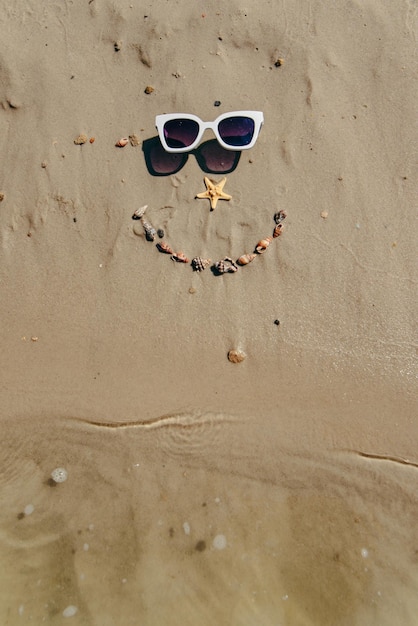 Faccina sorridente disegnata sulla sabbia con conchiglie e occhiali da sole vista dall'alto