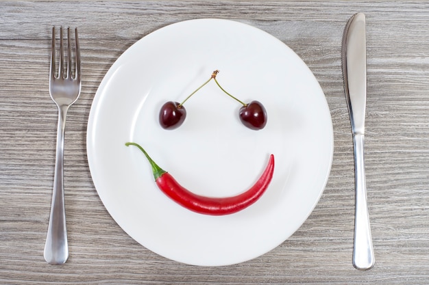 Faccina sorridente fatta di ciliegia e peperoncino su piatto con forchetta e coltello