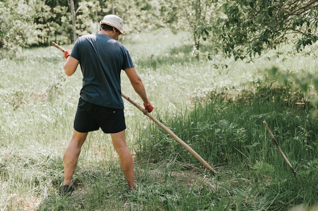 Falciare l'erba tradizionale vecchio stile con la falce a mano nella fattoria del villaggio domestico giovane contadino maturo che falcia l'erba verde erba cresciuta di un terreno agricolo con una falce