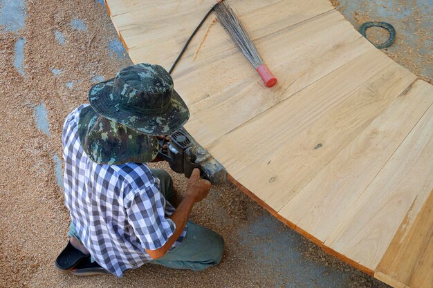 Falegname asiatico che utilizza una pialla elettrica per radere il bordo della panca di legno curva in un'officina all'aperto