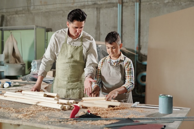 Falegname maschio in grembiule che indica le assi mentre spiega al figlio adolescente come lavorare con il legno nel laboratorio di mobili