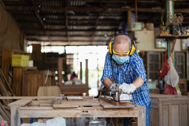 Falegname senior asiatico indossare maschera igienica proteggere utilizzando pialla elettrica regolare la superficie su tavola di legno presso il laboratorio di falegnameria