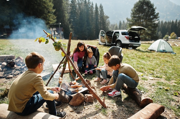 Falò di famiglia in montagna. Salsicce fritte. Madre con quattro bambini in campeggio. Escursione autunnale e clima del campo. Riscaldare e cuocere vicino alla fiamma insieme.