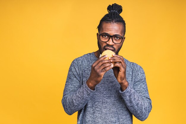 Fame giovane americano africano uomo nero mangiare hamburger isolate su sfondo giallo. Concetto di dieta.
