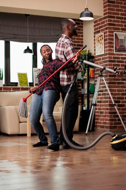 Famiglia afroamericana che fa lavori domestici in appartamento urbano, suonando musicista con scopa e aspirapolvere. Coppia sposata entusiasta che prova gioia mentre fa le faccende domestiche.