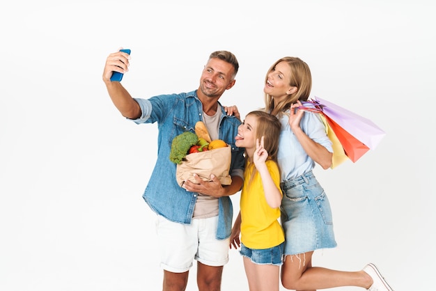 Famiglia allegra che indossa abiti casual in piedi isolata su bianco, facendo la spesa insieme, portando borse, facendo un selfie