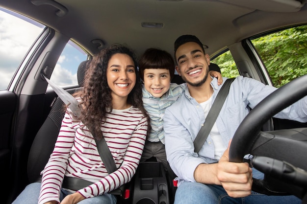 Famiglia araba felice con il piccolo figlio che si siede all'interno dell'auto e sorride alla macchina fotografica, genitori mediorientali allegri con bambino maschio sveglio che gode del viaggio nella loro nuova automobile, colpo del primo piano