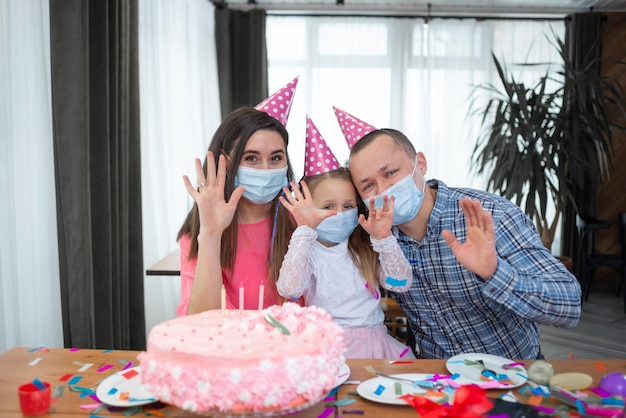 Famiglia caucasica che festeggia mentre si siede a casa al tavolo Maschere mediche e cappellini da festa che fanno un concetto di compleanno di videochiamata durante una pandemia