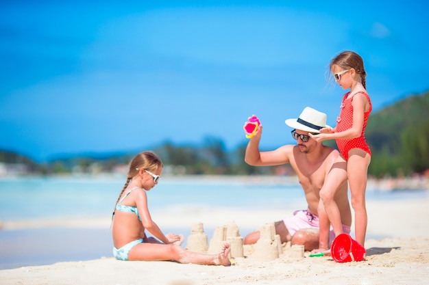 Famiglia che fa il castello della sabbia alla spiaggia bianca tropicale. Generi e due ragazze che giocano con la sabbia sulla spiaggia tropicale