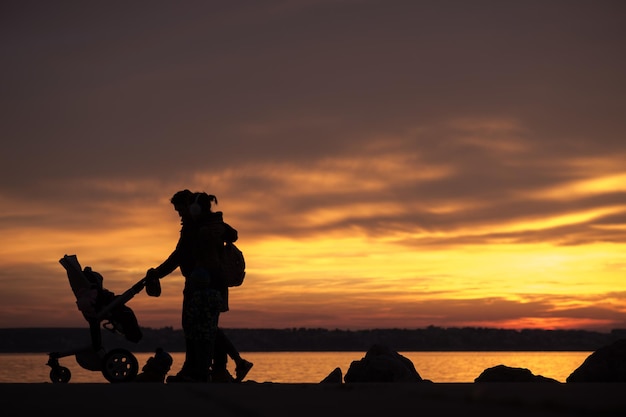 Famiglia con bambini e un bambino in carrozzina o passeggino che si staglia contro il tramonto arancione mentre fanno una passeggiata lungo la riva del mare.
