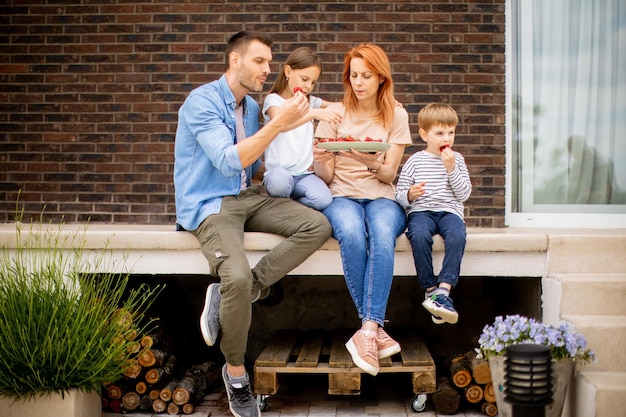 Famiglia con una madre padre figlio e figlia seduti fuori su un gradino di un portico anteriore di una casa di mattoni e mangiare fragole