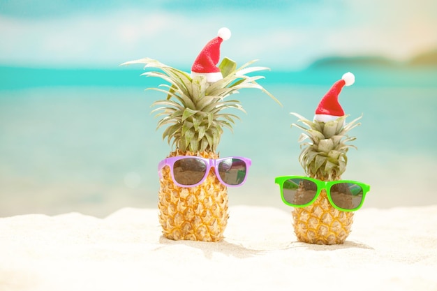 Famiglia di ananas divertenti e attraenti in eleganti occhiali da sole sulla sabbia contro il mare turchese. Indossare cappelli di Natale. Concetto di vacanza di Natale e Capodanno sulla spiaggia tropicale. Vacanza in famiglia. Luminoso