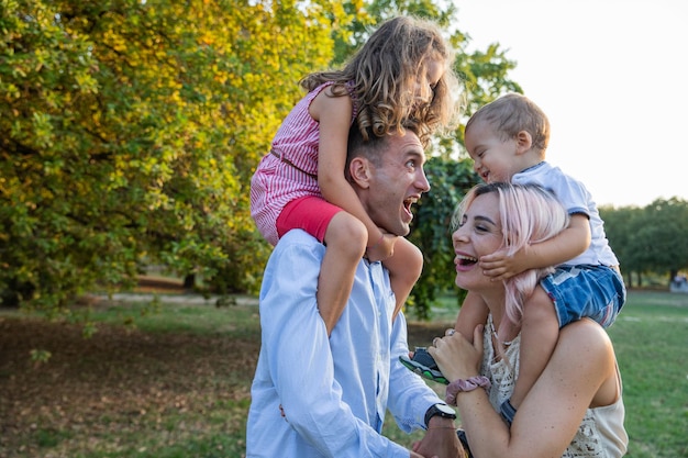 Famiglia di quattro persone al parco Bambini sulle spalle papà spalanca la bocca e fa ridere mamma e bambini