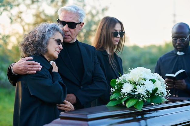 Famiglia di tre persone in lutto e sacerdote in abiti neri in piedi davanti alla bara