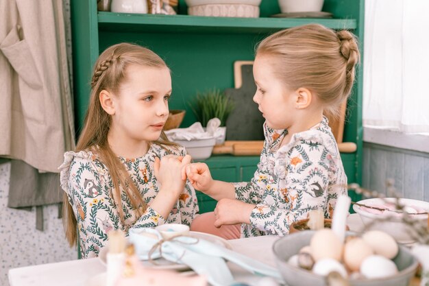 Famiglia felice candido bambini sorelle ragazze insieme giocoso mignolo giurano e si divertono pronti e aspettano le vacanze di Pasqua primaverili a casa in cucina tavola decorata per pranzo o cena