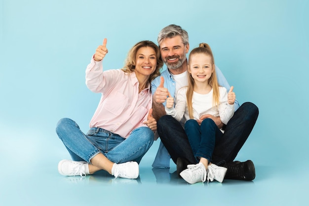 Famiglia felice che gesturing i pollici in su sorridente seduto su sfondo blu